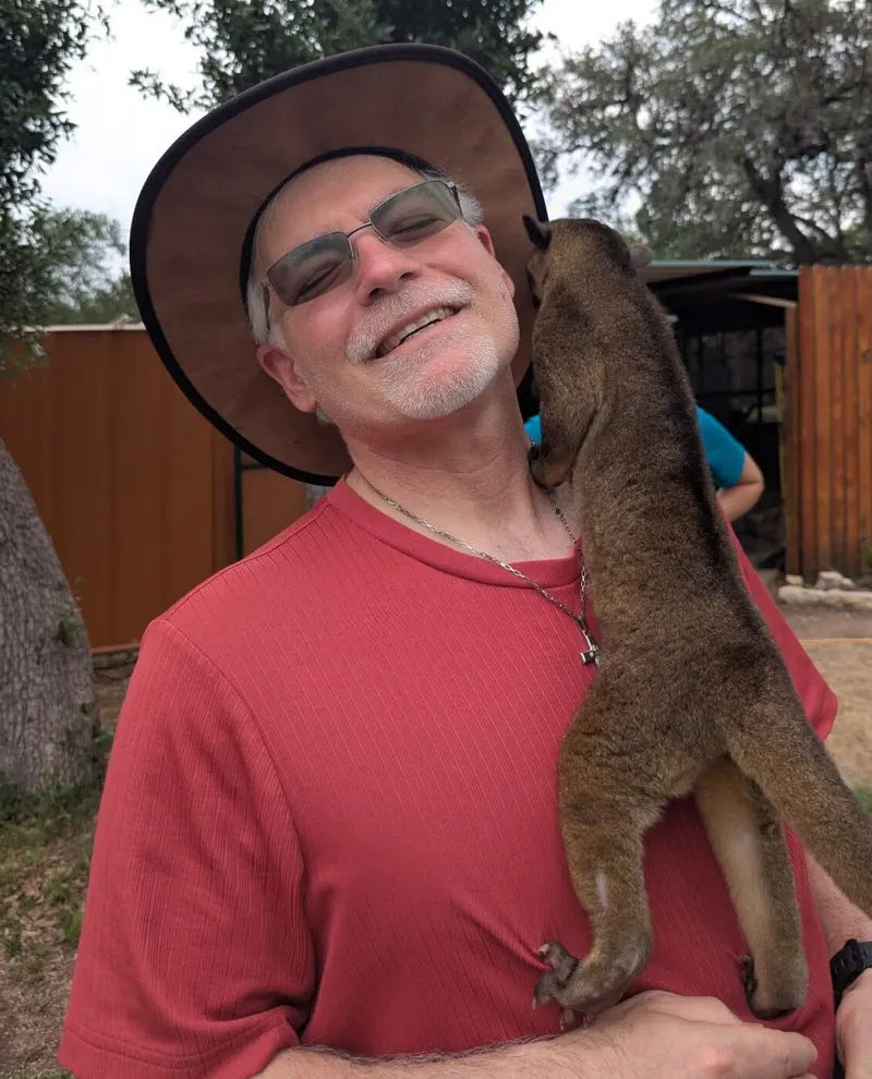 A handler in a sun hat smiles with Tulio the kinkajou perched on his shoulder.